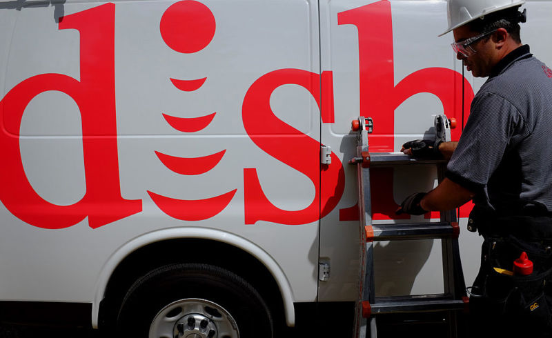 A technician in a hard hat stands next to a Dish Network service vehicle.